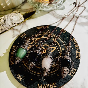 May include: Four crystal pendulums with decorative metalwork and chains are displayed on a black wooden board. The pendulums have green, purple, pink, and black gemstones. The board features gold text reading "YES", "MAYBE", and "REPHRASE", with geometric designs.