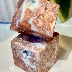 May include: Two stacked, cube-shaped decorative objects. The cubes are made of a stone material with a mix of colors, including shades of brown, white, and hints of blue. The stone has a unique, textured pattern. Each cube has a hollowed-out section.