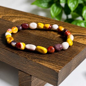 May include: A beaded bracelet featuring smooth, irregularly shaped stones in a variety of colours including mustard yellow, deep maroon, cream white, and burnt orange. The stones are strung together in a circular pattern to form a flexible wrist accessory.
