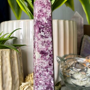 May include: A tall, rectangular crystal obelisk with a pointed top. The crystal is a mix of purple and white, with a speckled pattern. It is displayed on a black, round base. The background includes plants and other decorative items.