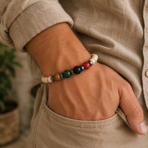 May include: A beaded bracelet featuring a sequence of round beads in cream, brown, green, black, and red, separated by small metallic spacers. The bracelet is worn on a wrist, with the beads arranged in a single strand around the arm.