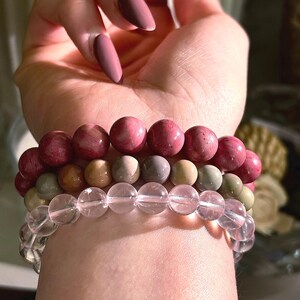 May include: Three beaded bracelets in shades of pink, grey, and clear. The top bracelet features large, round, pink beads. The middle bracelet has smaller, grey and tan beads. The bottom bracelet is made of clear, round beads.