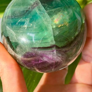 May include: A hand holds a polished fluorite sphere. The crystal ball features a mix of green, purple, and clear sections. The sphere has a smooth, reflective surface, with visible internal patterns and color variations. The overall effect is a vibrant, natural gemstone.