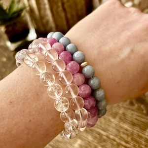 May include: Three beaded bracelets in clear, pink, and blue hues. The bracelets are made of round beads and are stacked on a wrist. The clear bracelet is at the bottom, followed by a pink and then a blue bracelet.