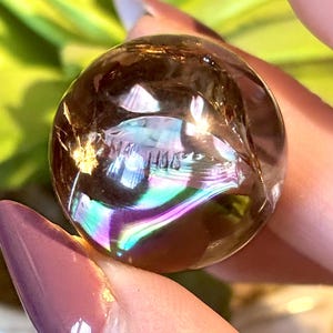 May include: A clear, iridescent crystal ball with a brown hue. The sphere reflects a rainbow of colors, including purple, blue, and green. The crystal is held between two fingers, with a blurred green background.