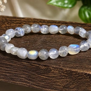 May include: A stretch bracelet composed of smooth, rounded labradorite beads. The beads are translucent with a greyish base color and exhibit iridescent flashes of blue and yellow light across their polished, rounded surfaces.