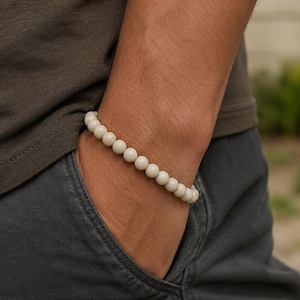 Puede incluir: Una pulsera elástica compuesta por cuentas redondas y lisas de color blanco roto. Las cuentas son uniformes en tamaño y forma, ensartadas en un bucle continuo que se ajusta perfectamente a la muñeca. Cada cuenta tiene un acabado mate.