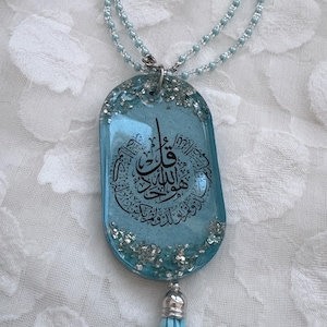 May include: A light blue pendant necklace with a silver chain and a matching tassel. The pendant is a rectangular shape with a light blue background and black Arabic calligraphy. Silver glitter accents the edges.