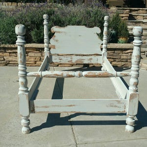 May include: A distressed, light blue bed frame with a headboard and four posts. The frame has a vintage, weathered appearance with visible paint chipping, revealing the wood underneath. The bed frame is designed for a standard mattress size.