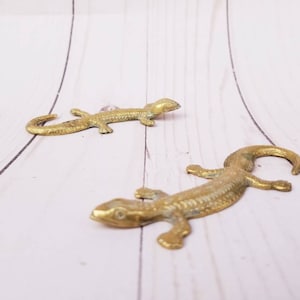 May include: Two gold-colored metal lizard figurines on a white wooden surface.