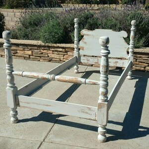 May include: A distressed, light blue bed frame with a headboard. The frame has four ornate posts and a footboard. The paint is chipped, revealing the wood underneath. The bed frame is empty and sits on a concrete surface.