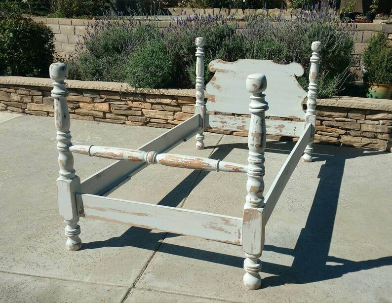 May include: A distressed, light blue bed frame with a headboard. The frame has four ornate posts and a footboard. The paint is chipped, revealing the wood underneath. The bed frame is empty and sits on a concrete surface.