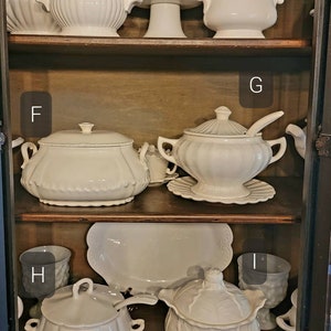 May include: A collection of white ceramic serving dishes and bowls with lids, some with decorative details, displayed on wooden shelves in a cabinet.
