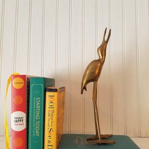 May include: A brass bird figurine standing on a stack of four books. The books are titled "Instant Happy Journal", "Starting Today", "1001 Things Your Kids Should See & Do", and "One Line A Day".