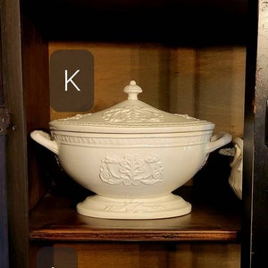 May include: Three white ceramic serving dishes with lids on a wooden shelf. The dishes are all different sizes and shapes. The top dish has a simple design, the middle dish has a floral design, and the bottom dish has a ribbed design.