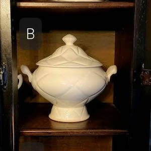May include: Three white ceramic serving bowls with lids are displayed on a wooden shelf. The bowls are different sizes and have different designs. The top bowl has a simple design, the middle bowl has a diamond pattern, and the bottom bowl has a more ornate design.
