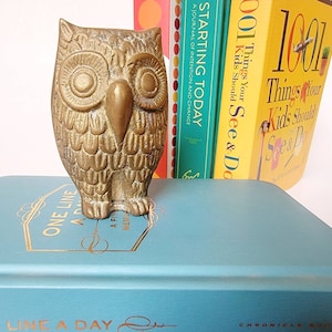 May include: A gold-colored brass owl figurine with detailed feathers and large eyes. The owl is sitting on top of a teal blue book with gold lettering that reads "One Line A Day" and "Chronicle Books".
