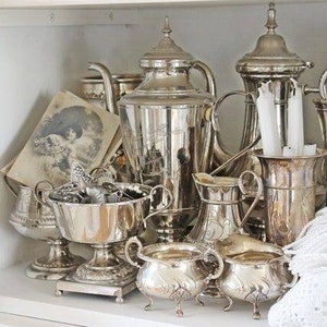 May include: A collection of silver-plated serving pieces, including a teapot, creamer, sugar bowl, and a vase, arranged on a white shelf.