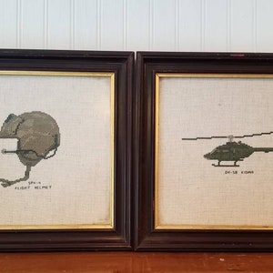 May include: Two framed cross-stitch pieces. The left piece shows a green military flight helmet with the text "SPHINX FLIGHT HELMET". The right piece shows a green helicopter with the text "UH-1B HUEY".