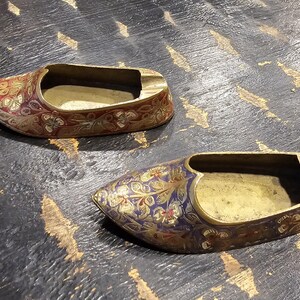 May include: Two ornate brass shoe-shaped ashtrays with intricate floral designs in shades of red, blue, and gold. The ashtrays are on a dark wooden surface.
