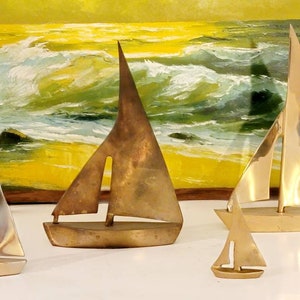 May include: Four brass sailboat figurines of varying sizes. The sailboats are arranged on a white surface in front of a painting of a seascape.