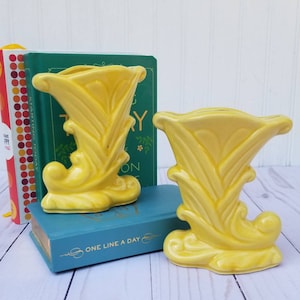 May include: Two yellow ceramic vases with a unique, ornate design. The vases are shaped like a stylized boot with a flared top. They are sitting on top of a blue book with the words "One Line A Day" on the cover.