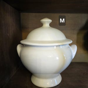 May include: White ceramic soup tureen with a lid. The tureen has two handles and a round base. The lid has a knob on top. The tureen is sitting on a wooden shelf. The letter "M" is visible in the upper right corner of the image.