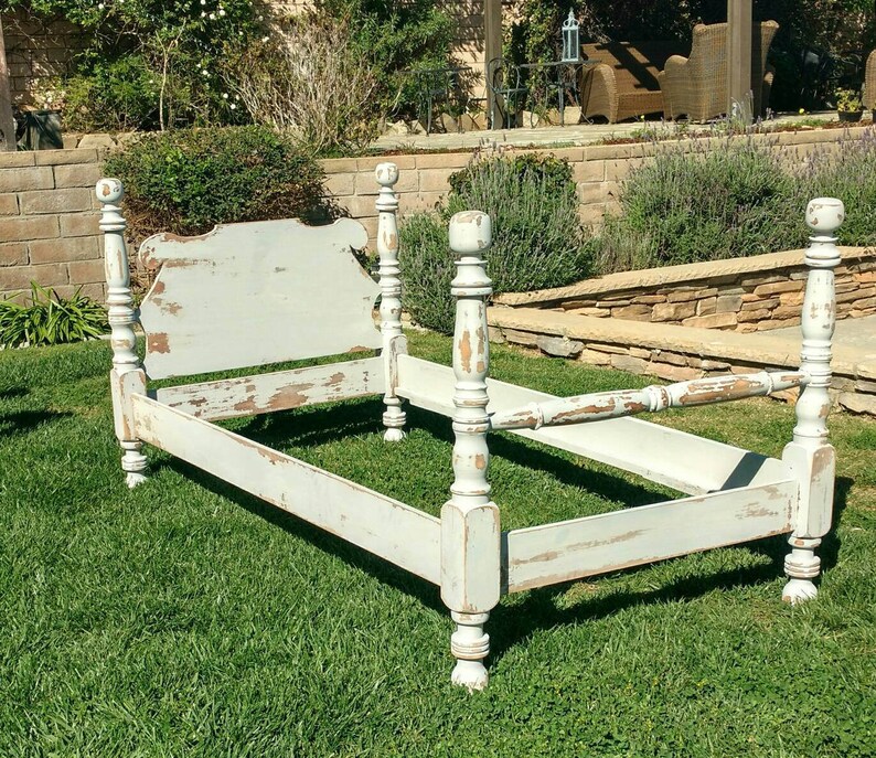 May include: A distressed, light blue bed frame with four tall, turned posts. The headboard has a decorative shape, and the paint is chipped, revealing the wood underneath. The bed frame is set on a green lawn.