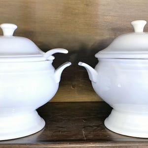 May include: Two white ceramic soup tureens with lids and ladles. The tureens are on a wooden shelf. The tureens are labeled "P" and "Q".