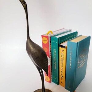 May include: A bronze crane ornament stands next to a stack of colourful books. The books have titles such as "Starting Today" and "One Line a Day." The crane is on a circular base.