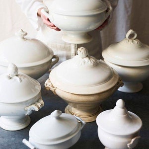 May include: A collection of six white ceramic serving bowls with lids. The bowls are all different sizes and have intricate details. The bowls are arranged on a gray surface.