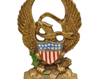 May include: A gold-colored cast iron doorstop shaped like an eagle with outstretched wings. The eagle is holding a shield with the American flag on it. The eagle is also holding a banner that says "E PLURIBUS UNUM" in a gold-colored ribbon. The doorstop is on a white background.