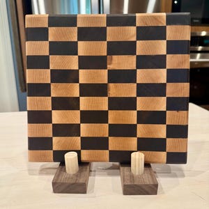 Peut inclure: Une planche à découper carrée en bois présentant un motif en damier composé de blocs de bois alternant entre le beige clair et le brun foncé. La planche est soutenue par deux supports en bois rectangulaires, chacun avec un tenon cylindrique.