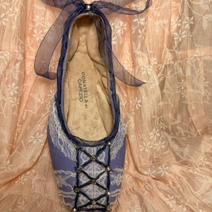 May include: A single lavender ballet slipper with white lace trim and a crisscross design on the toe. The slipper has a dark blue trim and a sheer ribbon bow. The interior is tan, and the brand "DONATELLA by CAPEZIO" is visible.