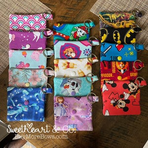 May include: A collection of small, fabric keychains with various patterns and character designs. The keychains feature designs like unicorns, cartoon characters, and floral prints. Each keychain has a button closure and a metal ring for attaching keys.