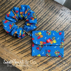 May include: A blue scrunchie and matching small wallet with a Rubik's Cube and "LOVE" pattern. The wallet has a red button closure and a silver clasp. The scrunchie and wallet are on a wooden surface.