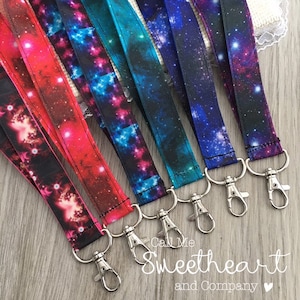 May include: A set of six galaxy-themed lanyards with silver swivel clips. The lanyards are made of black fabric with a galaxy print in shades of pink, blue, and purple.