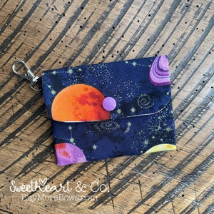 Pode incluir: Uma pequena bolsa de tecido azul escuro com fecho de correr e um chaveiro prateado. A bolsa tem um estampado com tema espacial com planetas, estrelas e constelações. Um botão roxo está preso à frente da bolsa.