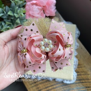 Puede incluir: Un lazo rosa con lunares dorados, detalles dorados y una pieza central de perlas y pedrería. El lazo está hecho de cinta en capas y tiene una malla dorada brillante. El texto "SweetHeart & Co. BuyMoreBows.com" es visible.