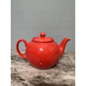 Könnte beinhalten: Eine leuchtend rote Keramik-Teekanne mit passendem Deckel und gebogenem Griff. Die Teekanne hat einen runden Körper und einen Ausguss zum Ausgießen. Der Deckel ist mit einem kleinen Knopf versehen. Klassisches Design.