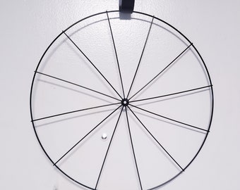 May include: A black metal wreath frame with a circular shape and spokes radiating from the center. The frame is hanging from a hook.