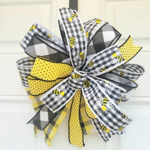 May include: A black and white checkered ribbon with yellow polka dots and a white ribbon with black and white checkered pattern and yellow bees printed on it. The ribbons are tied together in a bow.