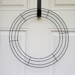 May include: A black metal wreath frame with a circular design, hanging on a white door. The frame is made of several concentric rings connected by vertical and horizontal bars, creating a grid-like structure.