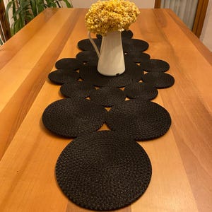 May include: A wooden table is set with a black woven table runner and matching coasters. A white pitcher holds a bouquet of yellow dried flowers. The table is made of light brown wood.