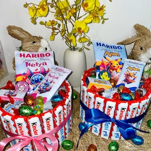 Peut inclure: Deux paniers de Pâques faits de barres chocolatées Kinder, remplis de bonbons et de friandises. Chaque panier est noué avec un ruban et entouré d'œufs en chocolat emballés dans du papier d'aluminium. Un vase de fleurs jaunes et des lapins décoratifs complètent la scène.