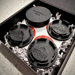 May include: A black box containing several black alignment tools. One tool is round with the text "ALIGNMENT TOOL". Three other tools have the text "stlsae". The tools are on white paper and red adhesive.