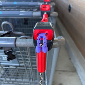 May include: A close-up of a shopping cart lock mechanism. The mechanism is red and black with purple key-shaped attachments. A silver chain hangs from the bottom. The cart handle is gray.