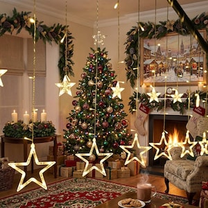 May include: A set of hanging LED star lights with warm white illumination, suspended from thin wires. The stars feature a clear frame and are arranged in a row, creating a festive display in front of a decorated Christmas tree and fireplace.