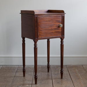 Może przedstawiać: Mały, antyczny stolik nocny z ciemnobrązowego drewna. Posiada pojedyncze drzwiczki z mosiężną gałką i stoi na czterech smukłych, toczonych nogach. Blat ma niewielki okap. Wysokość ok. 76 cm.