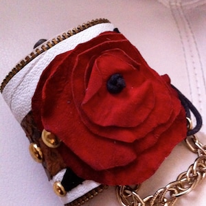 May include: A white and brown leather cuff bracelet with a large red fabric rose and gold chain accents.